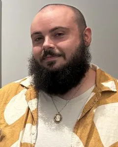 Image of Willett. Willett has short black hair and a beard. Willett is wearing a yellow and white patterned button up shirt over a white shirt.