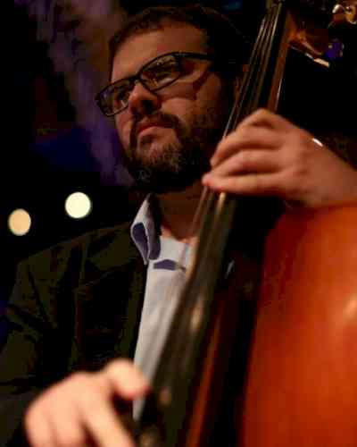 Photo of Rafael Abdalla playing the bass. Rafael has short brown hair, a beard, and black framed glasses. He is wearing a black suit.