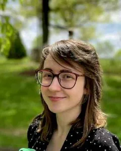 Photo of Anna Higgins. Anna has red glasses, medium length brown hair, and a black shirt with polka dots.