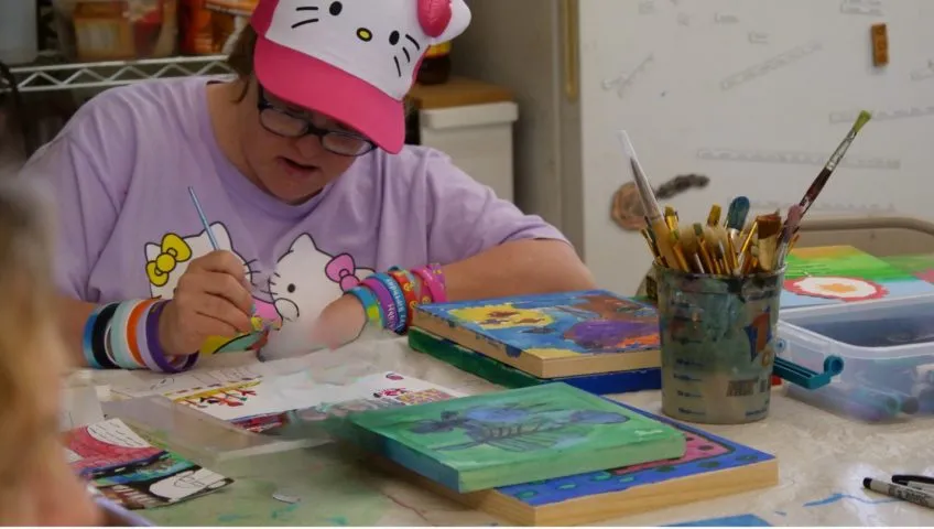 A person painting on a table surrounded by other paintings with a cup full of paint brushes nearby.