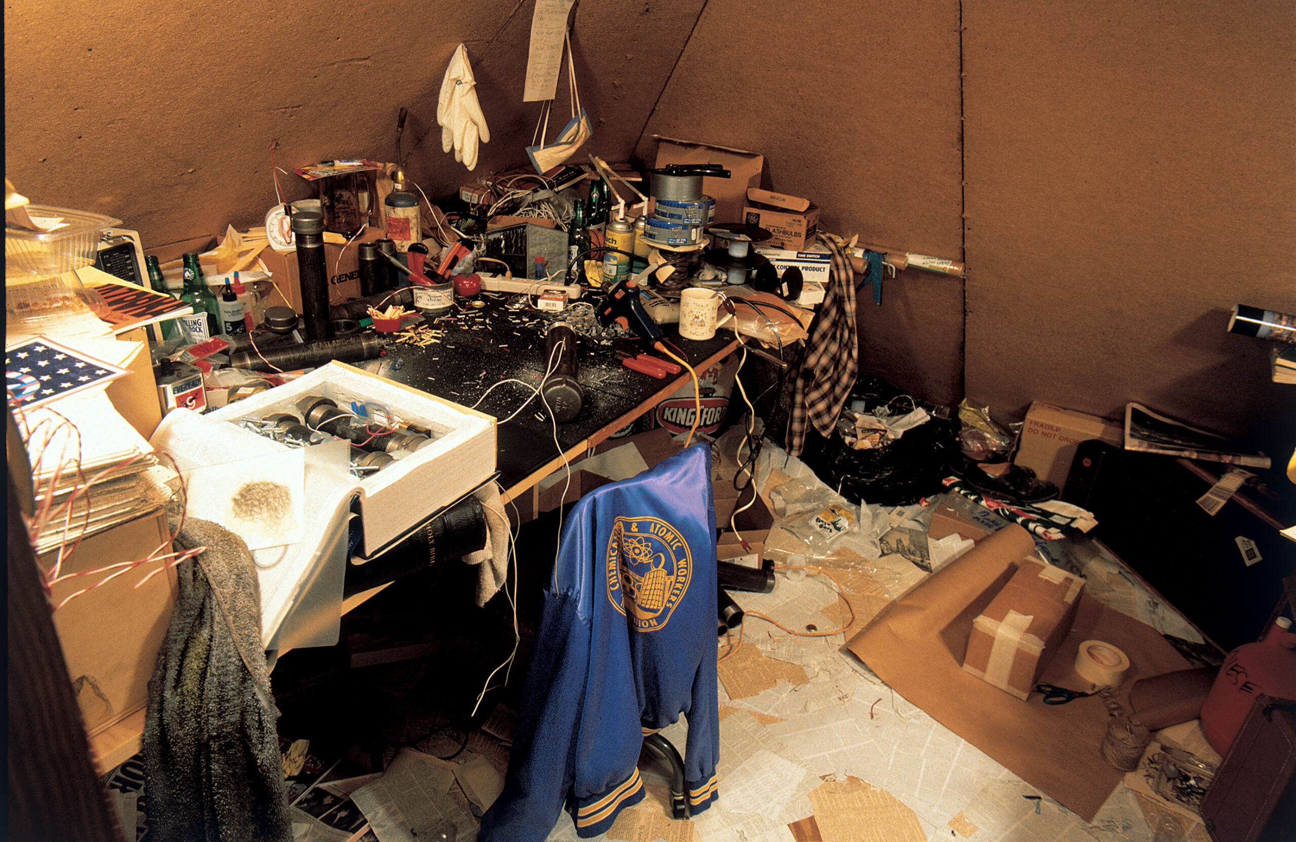 Photo of installation consisting of a hidden room that has a cluttered workstation with materials to create bombs are placed.