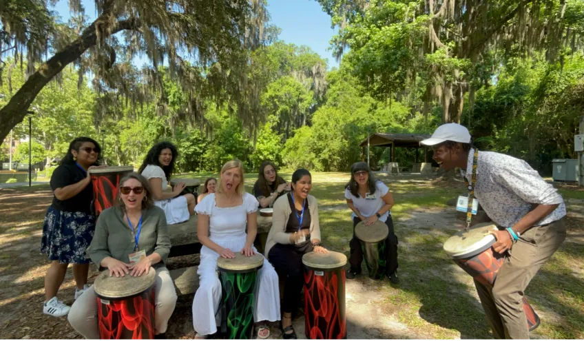 A group of people playing drums outside under green oak trees.