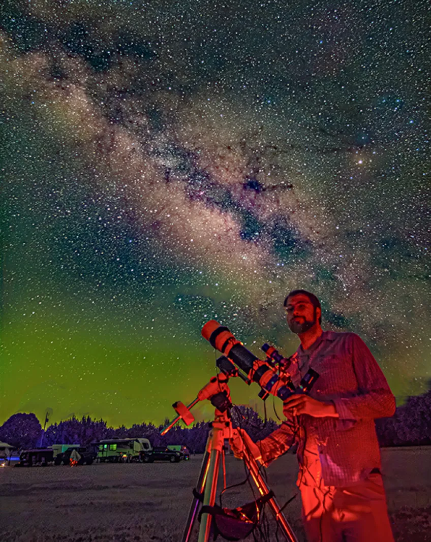 A person poses with a telescope at a dark sky site with the Milky Way clearly visible overhead.