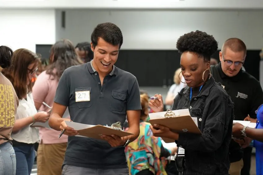 Two people holding clipboards and smiling at each other in a crowded room of attendees.