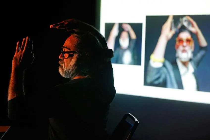 A person with a beard, wearing glasses, gestures with his hands under a spotlight. Behind him, two similar images are projected on a screen, reflecting his pose.