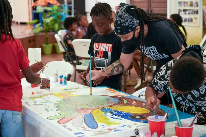 Group of children and an adult engage in a colorful painting project, sharing brushes and creativity. The atmosphere is lively and collaborative.