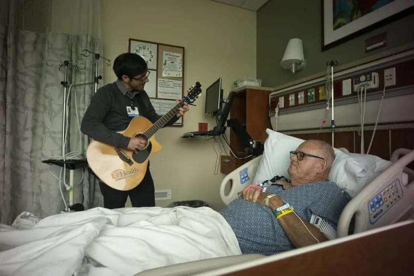 A musician plays guitar for a patient in a hospital bed. Medical equipment surrounds the bed and musician.