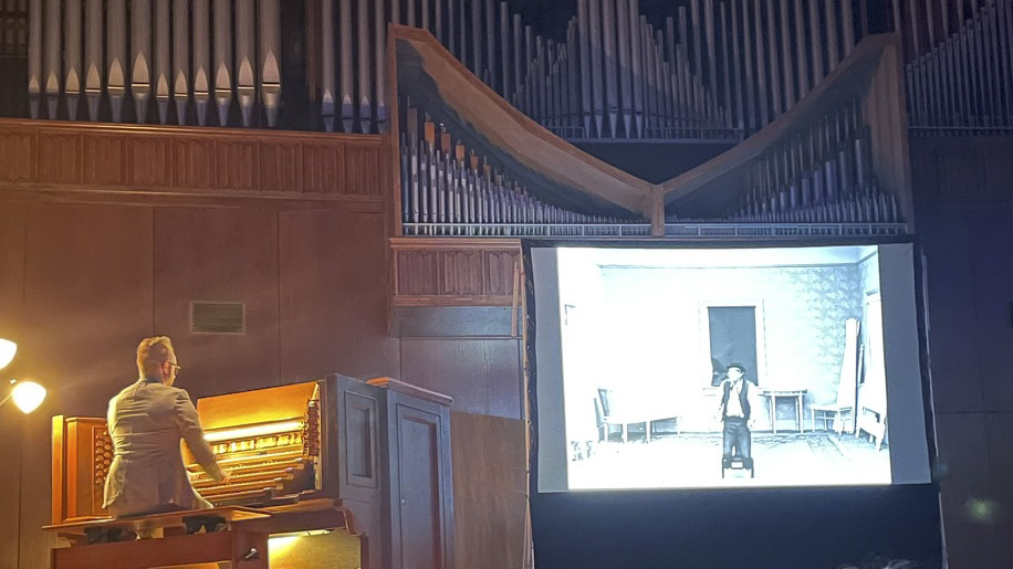 A person plays a pipe organ with an early 20th century silent film projected next to the organ.