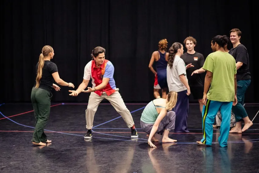 A group of people in casual attire rehearsing a scene on a stage with a black curtain backdrop.