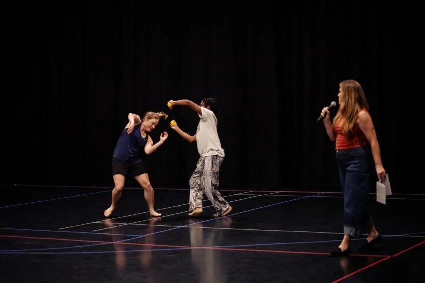 Two dancers perform, one holding tennis balls, on black stage while a woman with a microphone watches from the side.