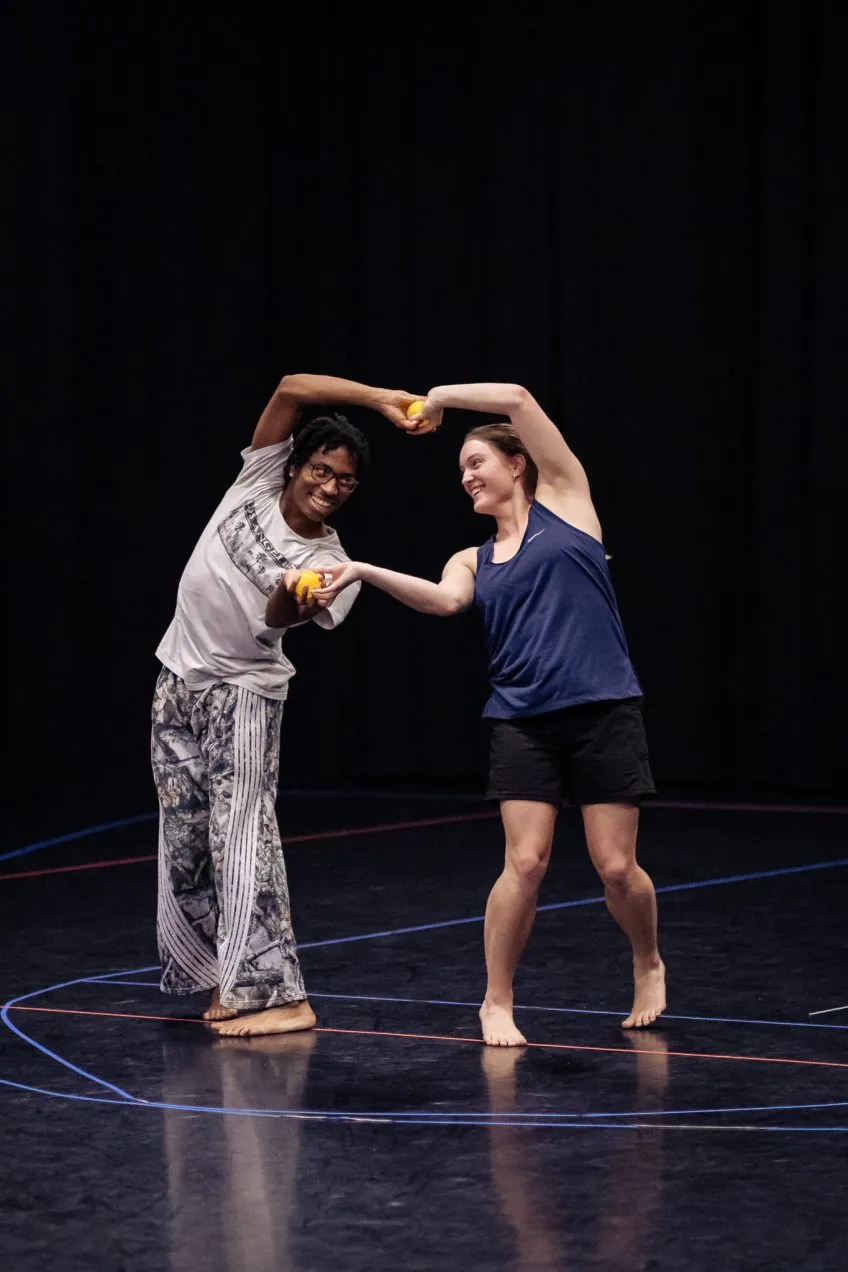 Two people dancing barefoot on a black stage with colored lines, holding hands and smiling.