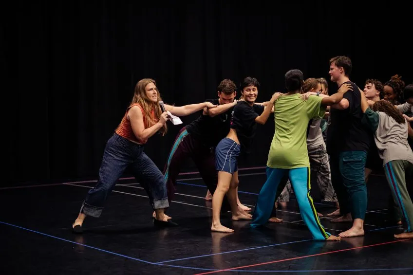 Dancers in a circle holding shoulders while a woman speaks into a microphone while gripping pulling one dancer's arm.