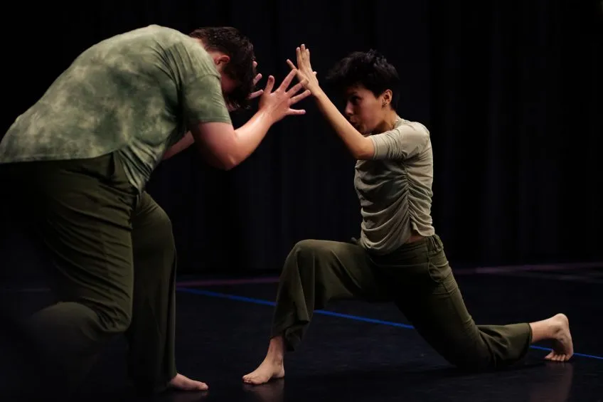 Two dancers in olive green clothing face each other with raised hands in a dramatic pose against a black curtain.