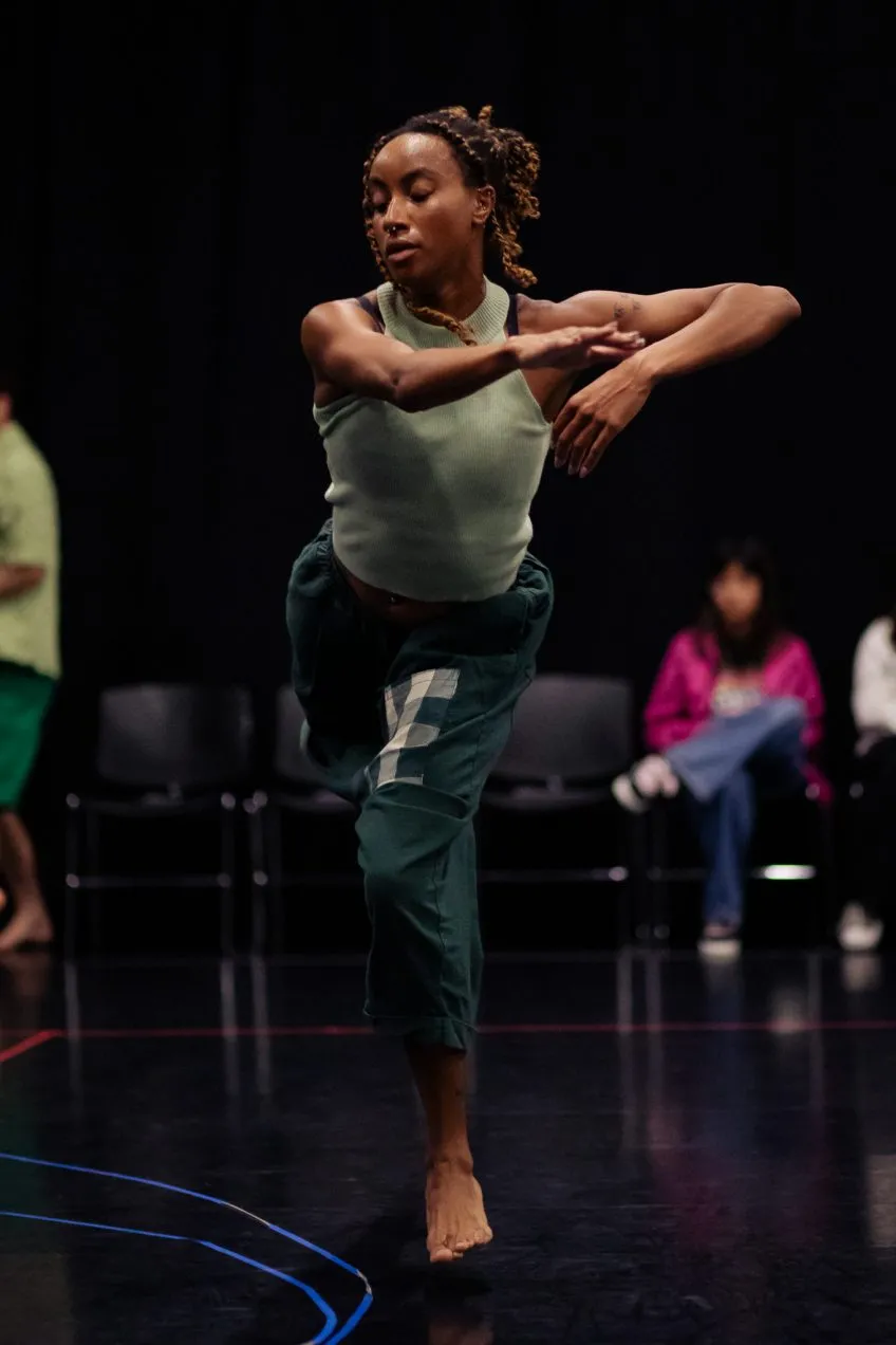 A woman in a green tank top and pants performs a dance move on a dark stage with others in the background.
