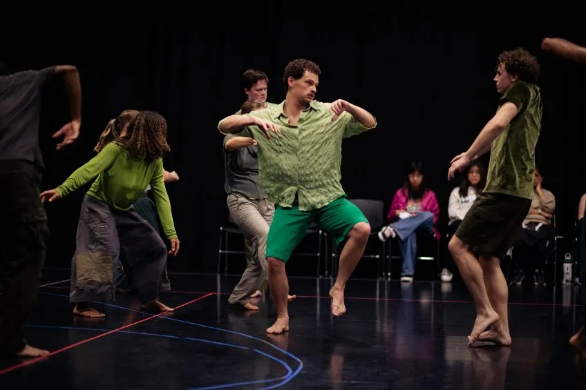 Group of people dancing barefoot on a dark stage with a seated audience in the background.