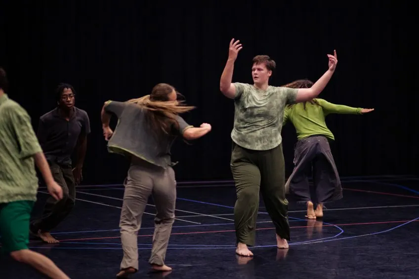Four people in casual clothing dance on a dark stage with thin lines marked on the floor.