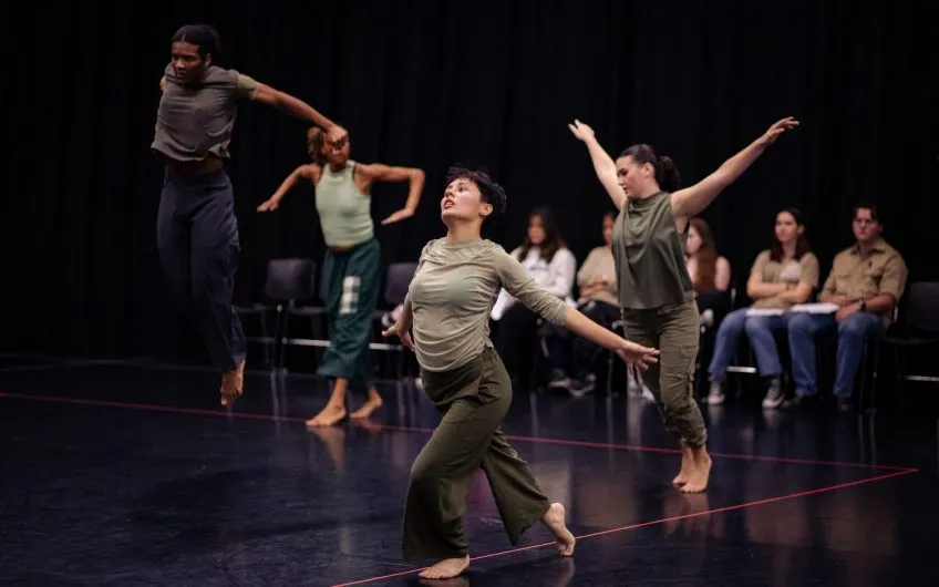 Four dancers in olive green attire performing contemporary movements on a black stage in front of a seated audience.