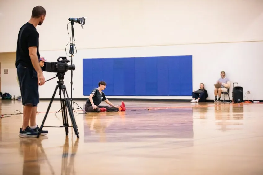 A videographer films a person stretching on a gym floor, with lighting equipment and crew in the background.