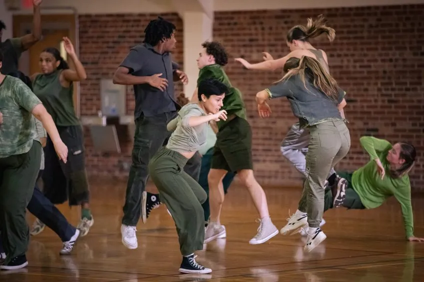 A group of dancers in olive green and grey athletic wear performing dynamic movements in a dance studio.
