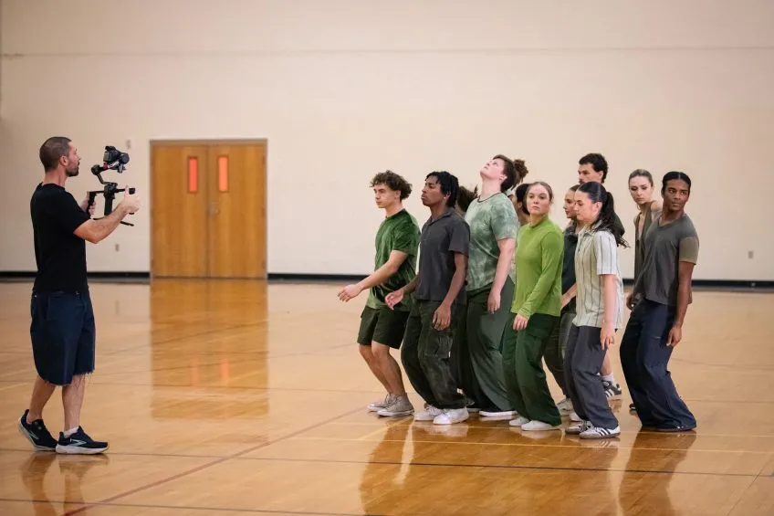 A videographer filming a line of seven people dressed in earthy-toned clothing in a gymnasium.