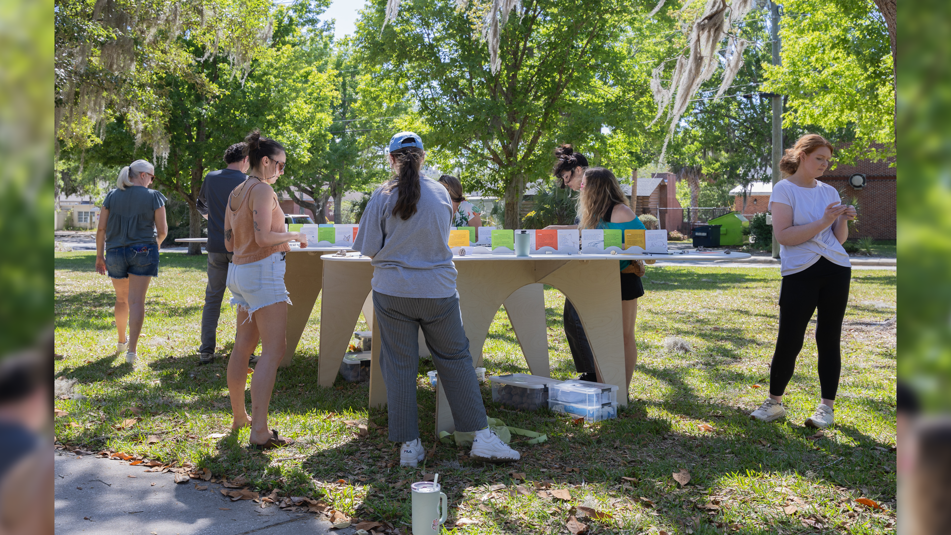 Students gather around a cardboard table displaying projects in a grassy outdoor area with trees.