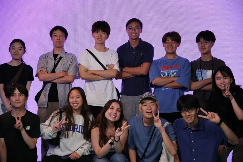 A group of twelve young adults posing for a photo against a bright, purple-lit grid background.