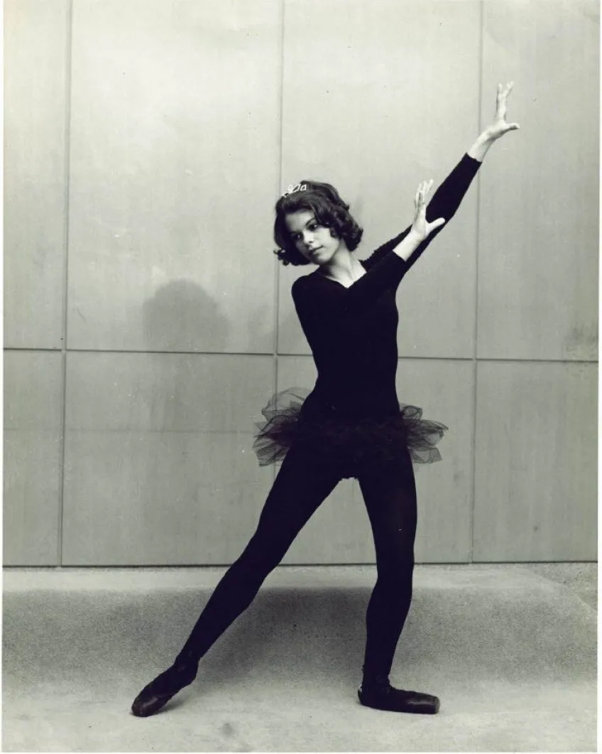 A ballerina in a black leotard and tutu posing with one arm raised against a paneled wall.