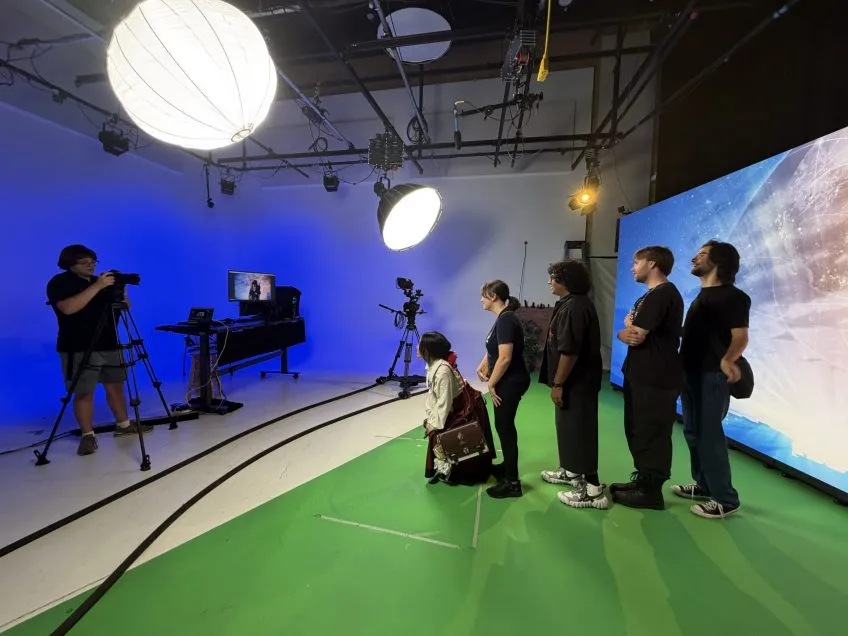 Film crew in a studio with a green screen floor, blue backdrop, large LED screen, and professional lighting.