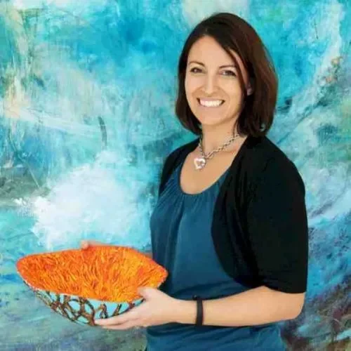 Heather Spooner smiling and holding a colorful, textured decorative bowl against an abstract blue background.
