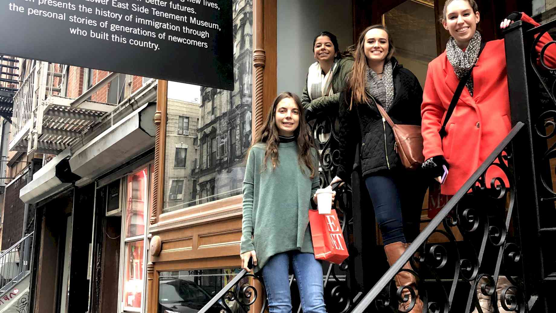 Museum Studies students on the stairs of the Tenement Museum in New York City. [Alt text: Four women pose on a wrought-iron staircase under a sign that reads “97 Orchard Street.”]