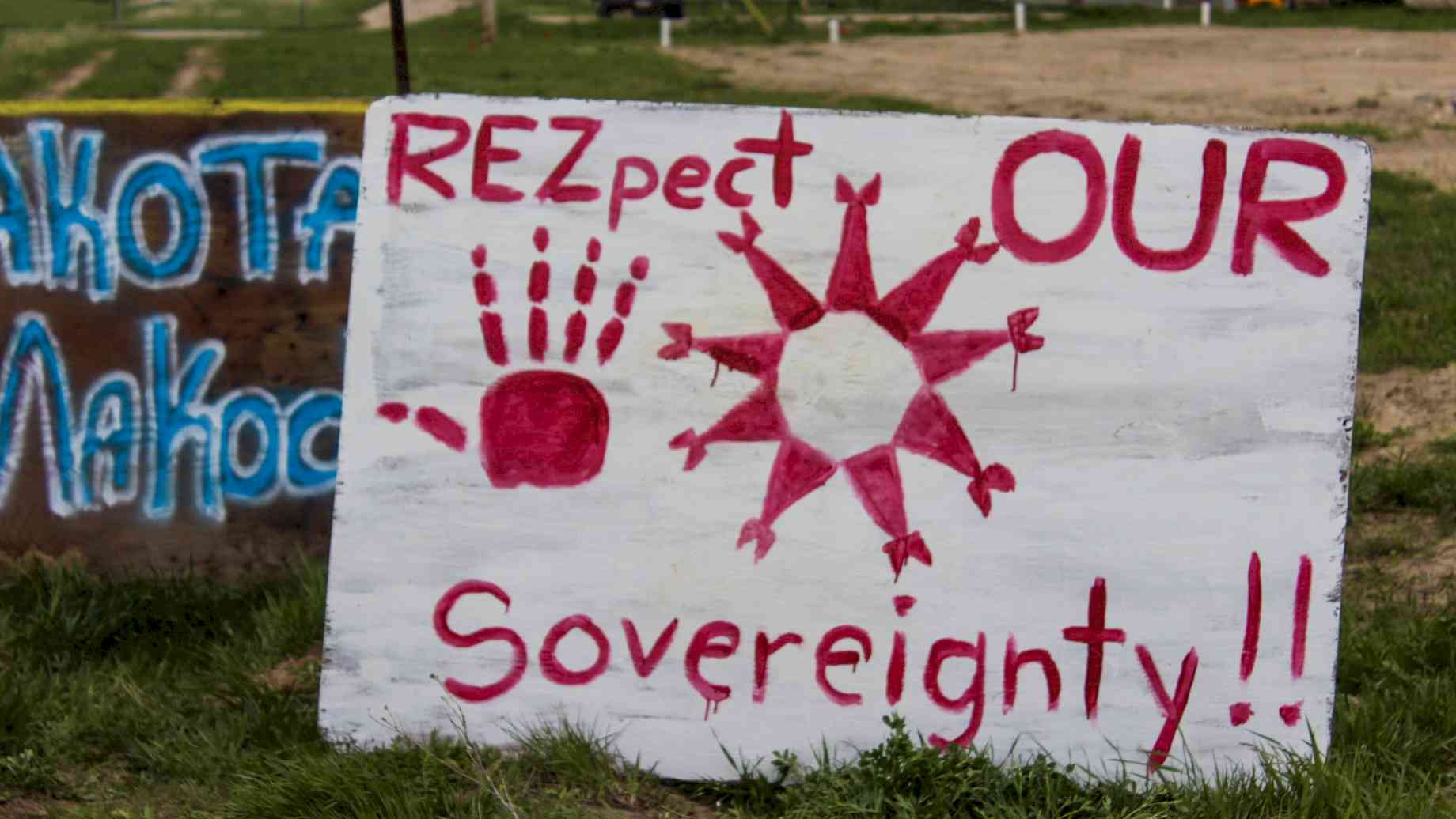 #respectoursovereignty. A piece of wood painted white with red lettering reads "R-E-Zpect our Sovereignty!" with a painted red hand facing palm out, and a circle of red triangles, pointing outward and topped with two additional triangles at the end of each point, facing inward like the letter "M." Credit: Pine Ridge Reservation