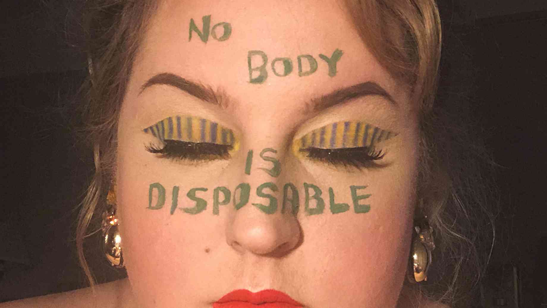 A woman with bare shoulders, a bolo necklace and gold earrings faces the camera with eyes closed. On her face in gold and green writing are the words "No Body is Disposable." Her eyelids are striped in the same colors.