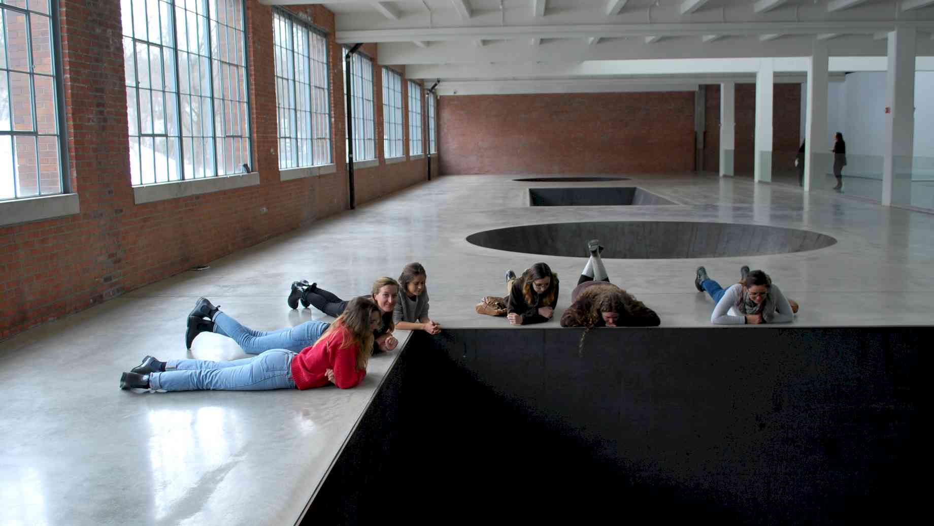 Museum Studies students experience art on a study trip to Dia: Beacon. [Alt text: Six students lie on the floor of a gallery, staring down into a black, square hole in the floor.]