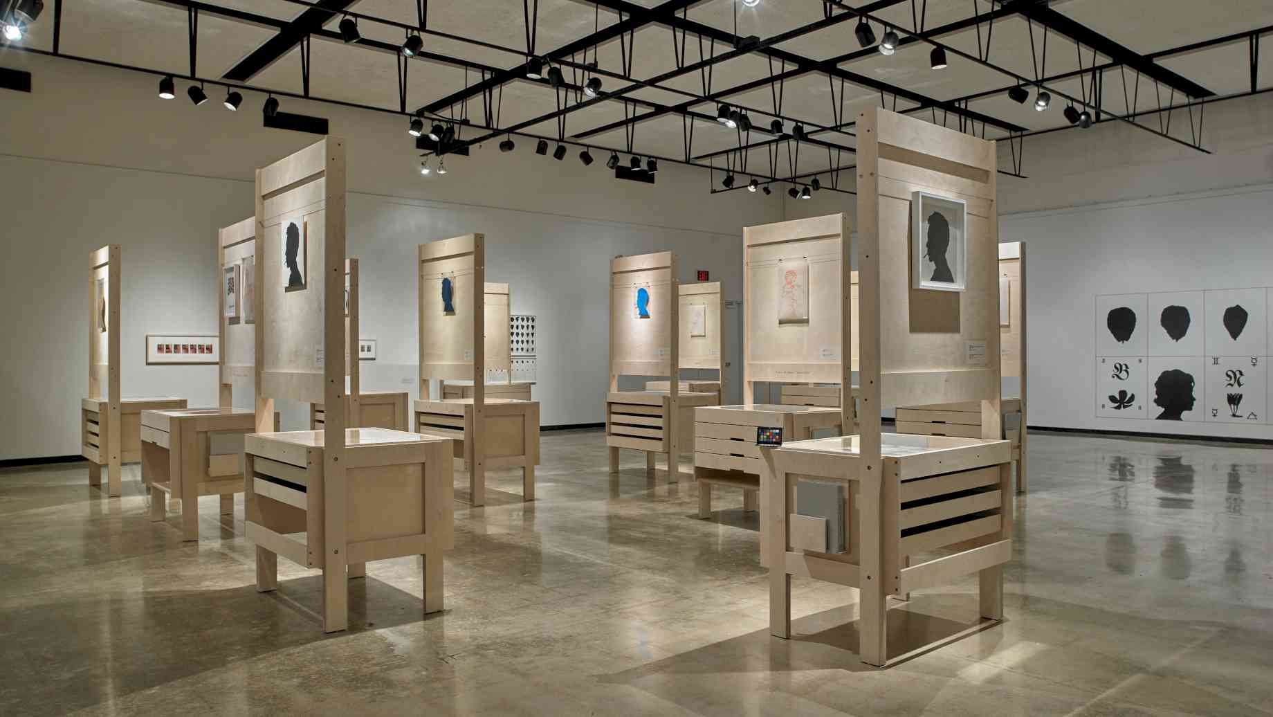 Exhibition in the University Gallery. Photo credit: UF University Galleries. [Alt text: Wooden desks are arranged in a large room, displaying silhouettes of faces.]